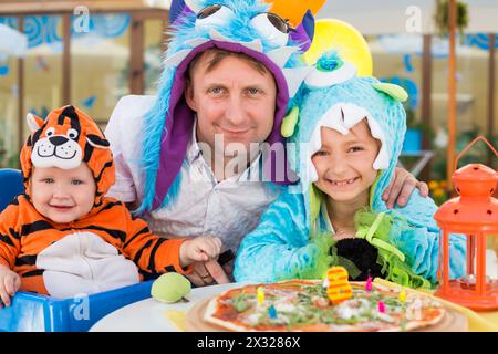 Padre con figlia in costumi da mostro e bambino in costume da tigre festeggiano il compleanno in un bar Foto Stock