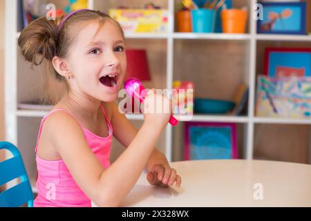 Bambina che canta in un microfono giocattolo seduto a un tavolo nella sala giochi Foto Stock