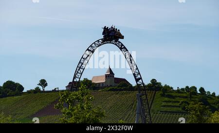 Le persone si divertono sul punto più alto delle montagne russe "Karacho" presso il parco a tema "Tripsdrill" con vigneti e una chiesa sullo sfondo Foto Stock