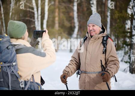 Foto media di un uomo caucasico anziano gioioso che indossa uno zaino in posa per la fotocamera mentre sua moglie scatta foto sullo smartphone Foto Stock