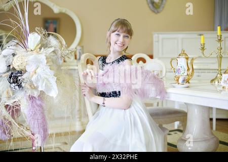 Una bellissima ragazza sorridente si siede a tavola con una serie di piatti e tiene in mano piume di struzzo. Foto Stock