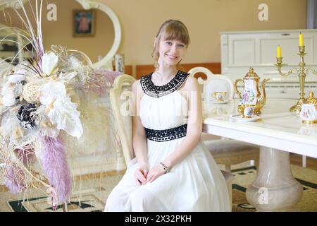 Una bellissima ragazza sorridente in abito bianco si siede a tavola con una serie di piatti. Foto Stock