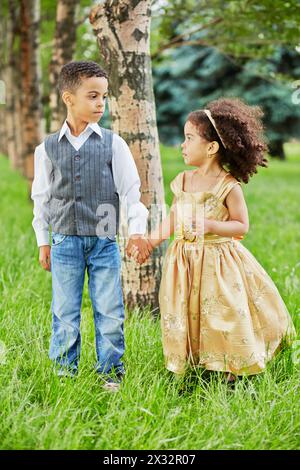 Bambina in beige da festa e ragazzo in piedi tenendosi per mano nel parco su un prato erboso Foto Stock