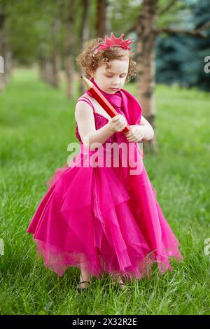 La bambina vestita con un luminoso abito gonfio esamina una grande matita rossa in mano in piedi su un prato erboso nel parco Foto Stock