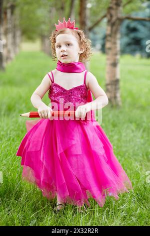 La bambina vestita con un luminoso abito gonfio si erge su un prato erboso nel parco tenendo in mano una grande matita rossa Foto Stock