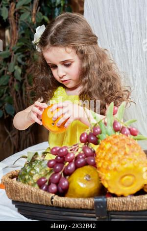 La bambina buccia il mandarino in piedi vicino a un cestino pieno di frutta Foto Stock