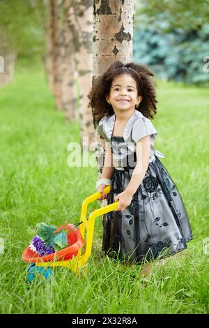 Una bambina sorridente vestita con un bellissimo abito nero si erge nel parco reggendo la carriola di plastica in cui giacciono i fiori Foto Stock