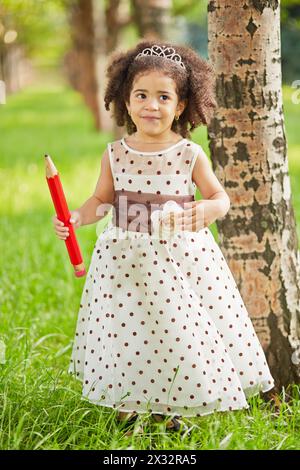 La bambina vestita con un abito a pois si trova nel parco tenendo una grande matita rossa in mano destra Foto Stock