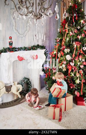 Un bambino si siede su una grande scatola regalo di cartone, tenendone un altro tra le mani vicino all'albero di Natale decorato, la bambina prende le decorazioni dalla confezione Foto Stock