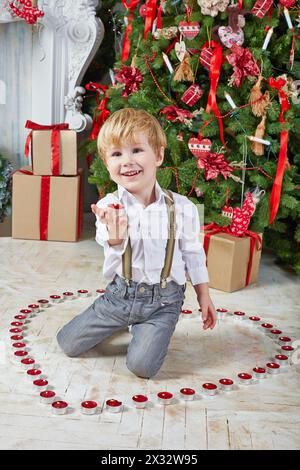 Un bambino si siede sul pavimento tra i tealights in fiamme e ne tiene uno sul palmo sotto l'albero di Natale Foto Stock