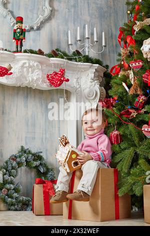 La bambina si siede su una scatola regalo di cartone con un aspetto appagato sotto l'albero di Natale, tenendo la casa del pan di zenzero nelle sue mani Foto Stock