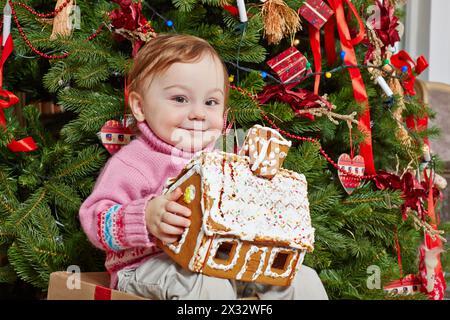 La bambina si siede su una grande scatola regalo di cartone con un aspetto appagante sotto l'albero di Natale, tenendo la casa del pan di zenzero nelle sue mani Foto Stock