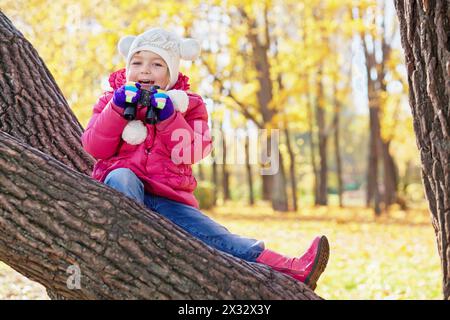 Una bambina con binoculare si siede sul tronco inclinato dell'albero nel parco autunnale Foto Stock