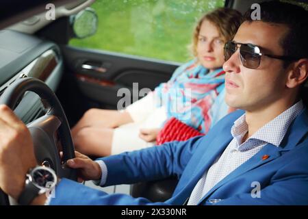 Un uomo con gli occhiali da sole guida un'auto e una donna si siede accanto a lui al giorno della pioggia. Concentrati sull'uomo. Foto Stock