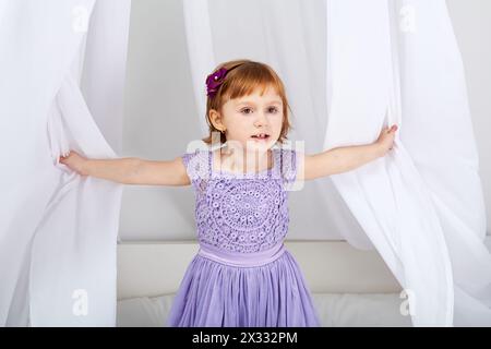 La bambina vestita con abiti da vacanza guarda fuori da sotto la tenda sul letto Foto Stock