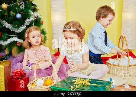 Tre bambini siedono nella stanza sotto l'albero di natale tra scatole regalo e cesti di vimini pieni di mandarini, fiocchi di neve artificiali e palle di neve Foto Stock