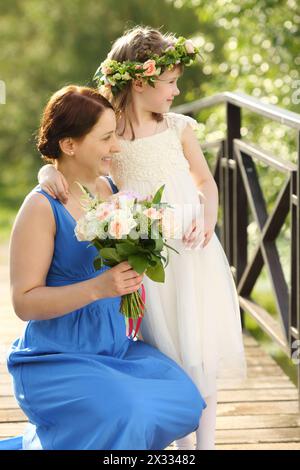 Bella madre con bouquet e piccola figlia in ghirlanda, guarda lontano sul ponte del parco. Concentrati sulle donne. Foto Stock