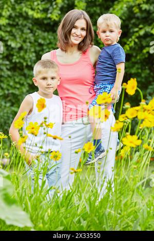 Una giovane donna sta in piedi con due figli piccoli sulla glade di fiori nel parco, tiene il figlio più giovane tra le braccia Foto Stock