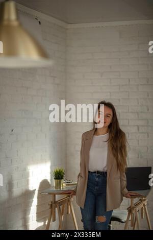 Giovane donna appoggiata a un tavolo con un portatile nelle vicinanze, inondata di luce naturale da una grande finestra. La scena suggerisce un lavoro produttivo Foto Stock