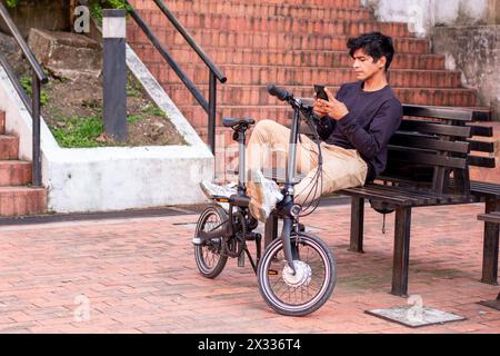 Stile di vita. Un giovane latino seduto accanto alla sua bicicletta elettrica e al suo cellulare in mano Foto Stock