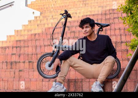 Un giovane Latino elegante che posa accanto alla sua bicicletta elettrica. seduti sulle scale in città Foto Stock
