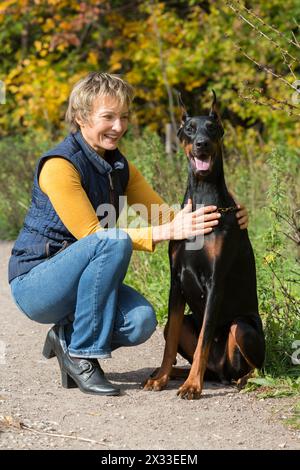 Una donna sorridente al collo di polo beige sta abbracciando dobermann in un parco. Foto Stock