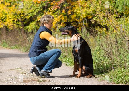 La donna al collo di polo beige è seduta di fronte al cane nero in un parco. Foto Stock