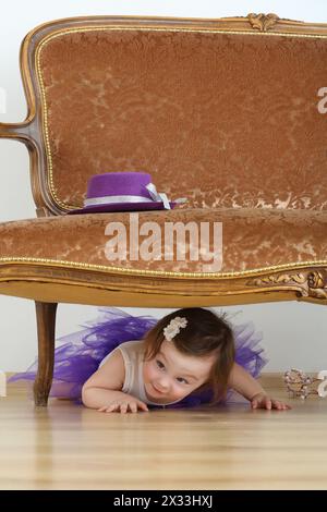 Bella bambina con una gonna viola stesa sul pavimento sotto il divano Foto Stock