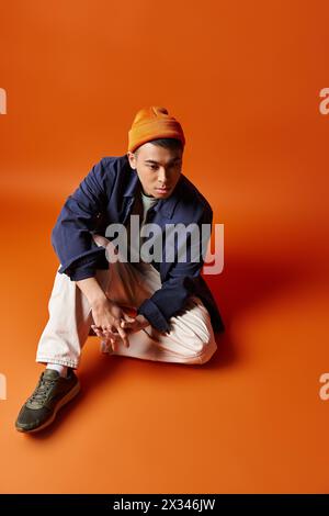 Un giovane uomo asiatico alla moda siede a terra, indossa un cappello alla moda, trasuda un'atmosfera contemplativa su sfondo arancione. Foto Stock