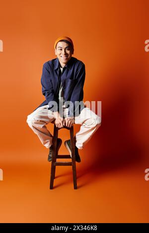 Un bell'uomo asiatico con un abbigliamento elegante siede graziosamente su uno sgabello di legno su uno sfondo arancione in uno studio. Foto Stock