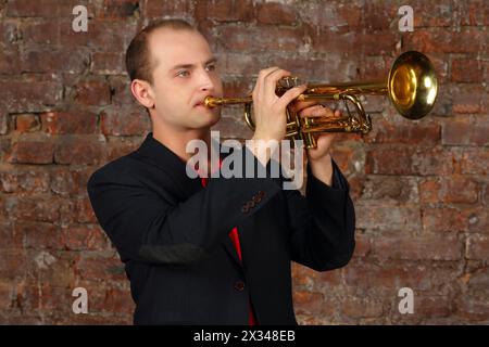 Il giovane bell'uomo in tuta suona la tromba in studio con il muro di mattoni Foto Stock