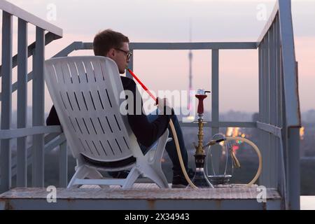 Il giovane fuma un narghilè seduto su una poltrona sul tetto alto, guardando il tramonto, vista posteriore. Foto Stock