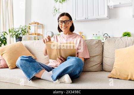 Una donna matura in intimi indumenti per la casa si rilassa su un divano, impegnata a leggere un libro. Foto Stock