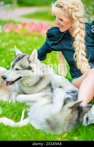 La giovane bionda gioca con due cani Husky seduti sull'erba nel parco estivo. Foto Stock