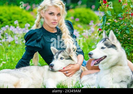 La giovane bionda gioca con due cani Husky seduti su un prato erboso nel parco estivo. Foto Stock