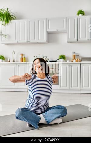 Una donna matura in intimo indumenti per la casa pratica yoga su un tappetino nella sua cucina. Foto Stock