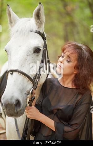 Donna in abito nero in piedi con cavallo bianco nel parco. Foto Stock
