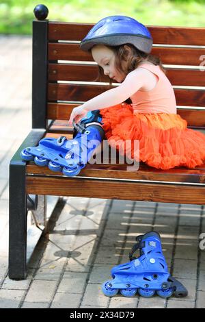 La bambina si siede sulla panchina e rimuove i pattini a rotelle blu durante il giorno estivo Foto Stock