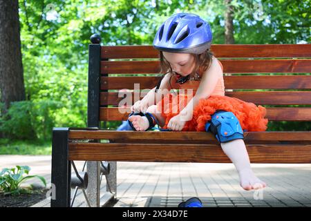 La bambina si siede sulla panchina e rimuove i cuscinetti protettivi per il pattinaggio a rotelle Foto Stock