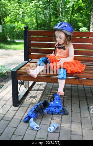 La bambina si siede sulla panchina e rimuove i pattini nel parco verde estivo Foto Stock