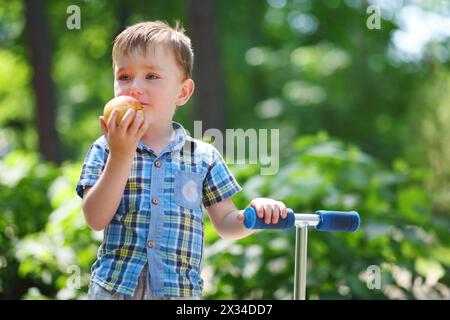 Il piccolo bel ragazzo mangia la mela vicino allo scooter nel parco estivo Foto Stock