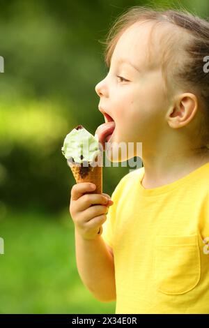 Una bambina carina in giallo lecca un gelato dolce nel verde del parco Foto Stock