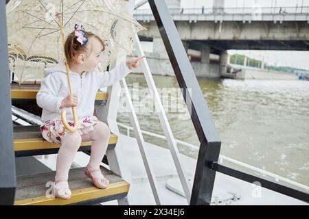 La bambina con l'ombrello si siede sul gradino sul ponte della barca e punta con le dita. Foto Stock