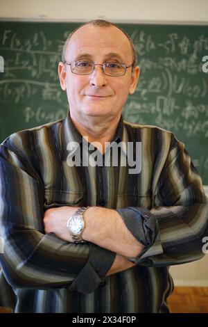 Un vecchio insegnante sorridente con le braccia incrociate si trova vicino alla lavagna con formule matematiche in classe scolastica Foto Stock