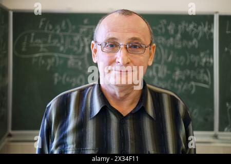 Un vecchio insegnante sorridente si trova vicino alla lavagna con formule matematiche in classe scolastica Foto Stock