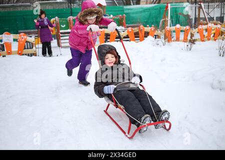 Ragazza felice che fa rotolare un bambino su una slitta di fronte alla ragazza con gli sci nel parco giochi invernale Foto Stock