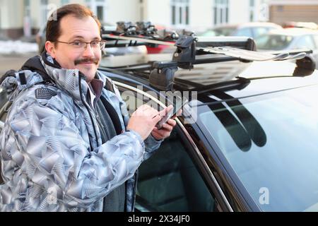 Uomo felice in occhiali con cellulare in mano vicino all'auto nel parcheggio Foto Stock