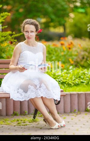 bella donna con gli occhiali, indossa un vestito bianco seduto sulla panchina e leggendo un libro, guardando la macchina fotografica Foto Stock