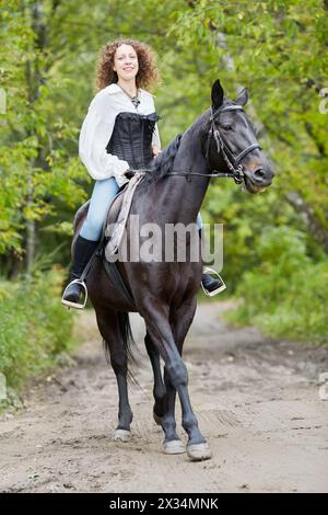 Donna sorridente con capelli ricci cavalcate sul cavallo da baia nel parco. Foto Stock
