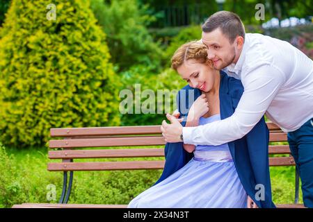 La donna si siede sulla panchina, l'uomo mette la giacca per ragazza nel verde del parco estivo Foto Stock